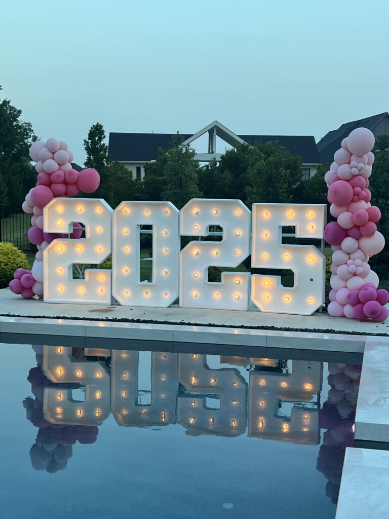 Brightly lit "2025" displays adorned with pink balloons beside a pool, creating a celebratory atmosphere for event.