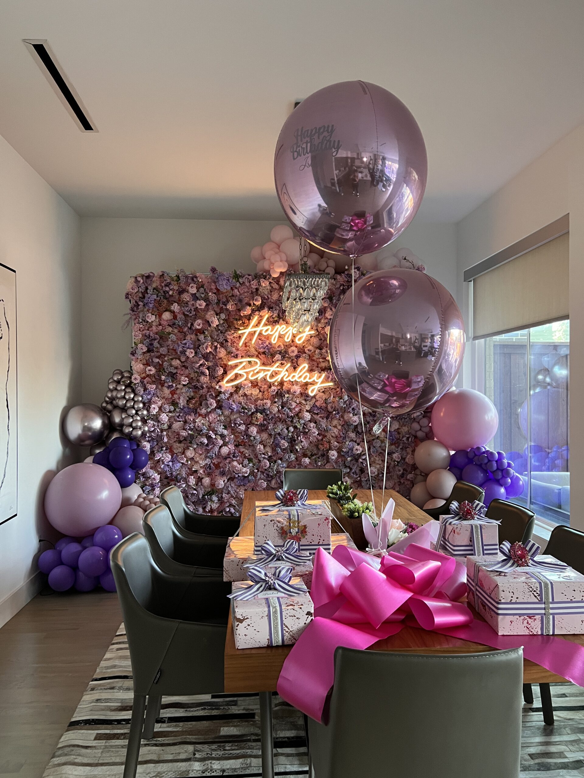 Colorful birthday celebration setup featuring a "Happy Birthday" neon sign, elegant décor, and wrapped gifts on a dining table.