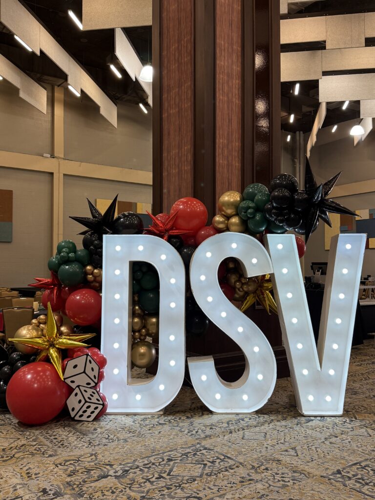 Illuminated letters "DSV" adorned with colorful balloons and decorative stars, set in a festive event space.