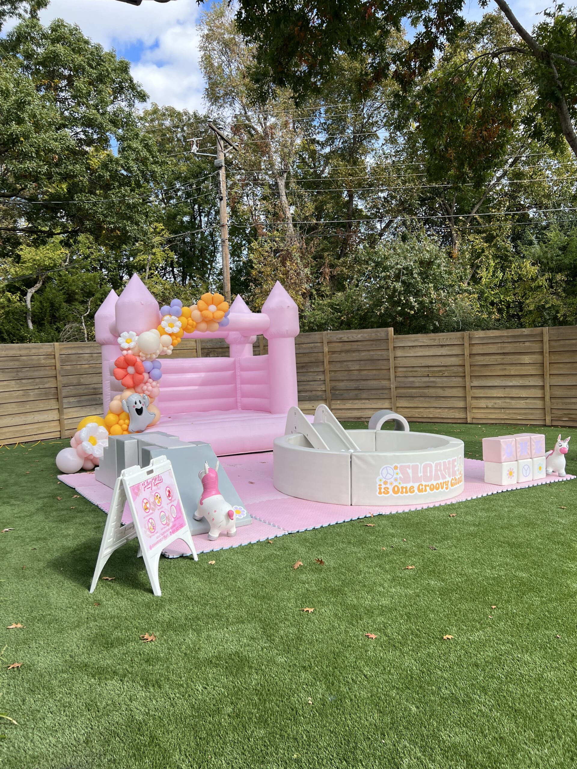 Bright pink bouncy castle surrounded by colorful balloons, a slide, and play areas on green grass