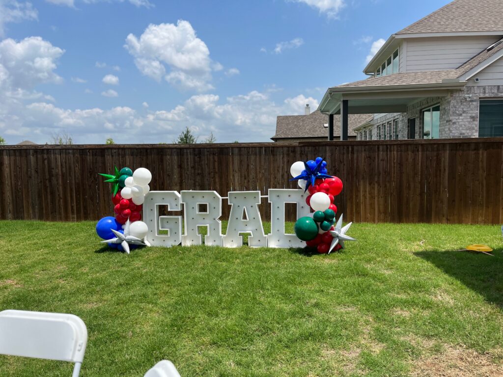 Large "GRAD" sign adorned with colorful balloons celebrates a graduation outdoors, set against a blue sky and wooden fence.