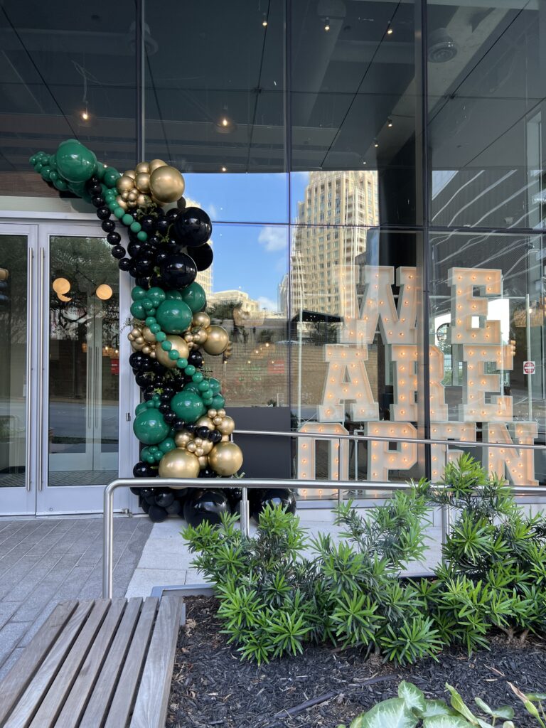 Colorful balloon arch in green, gold, and black decorates the entrance, complemented by illuminated letters stating "WE ARE OPEN."