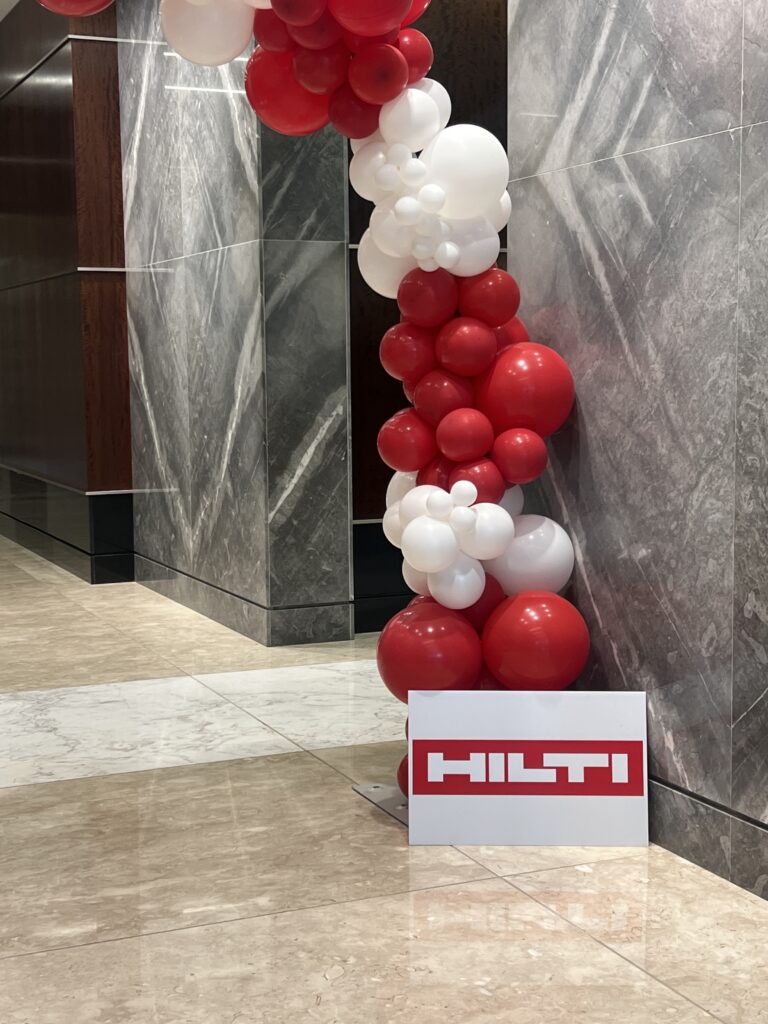 A festive balloon arch in red and white colors, featuring a prominently displayed Hilti sign, enhances the entrance of a modern building.