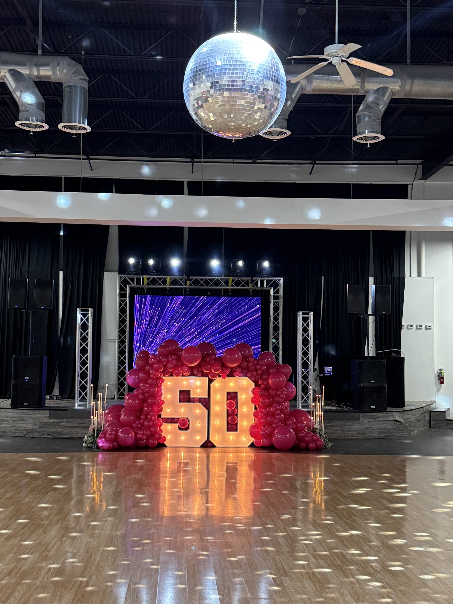 A festive décor featuring a large illuminated "50" surrounded by pink balloons, with a disco ball above, set for a 50th birthday celebration.