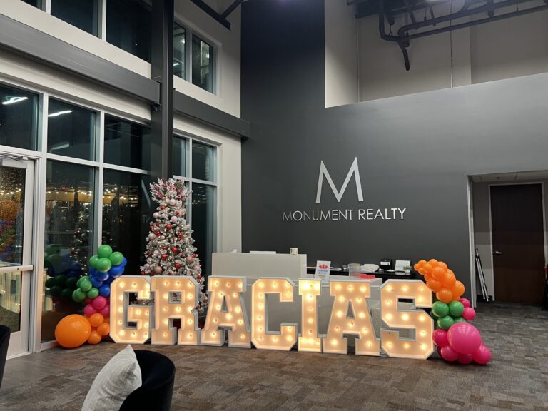 Illuminated "GRACIAS" sign in a modern office decorated for an event, featuring a Christmas tree and colorful balloons, reflecting a festive atmosphere.