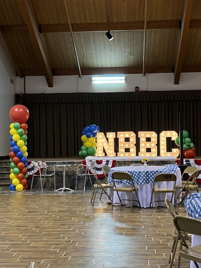 Brightly decorated event space featuring large illuminated "NBBC" letters, colorful balloon pillars, and checkered tablecloths, ideal for celebrations.