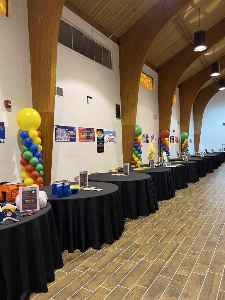 A spacious event hall features decorated tables with informational displays, colorful balloon columns, and warm wooden accents, showcasing community resources.