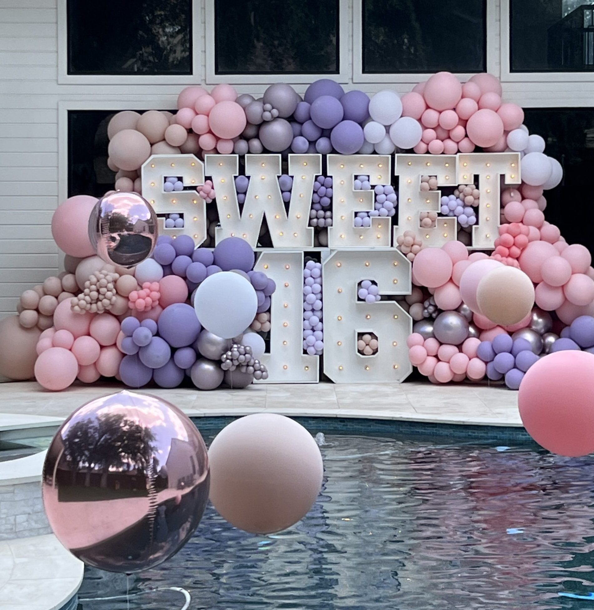 Colorful balloon decor showcases a lit "SWEET 16" sign, celebrating a milestone birthday near a pool