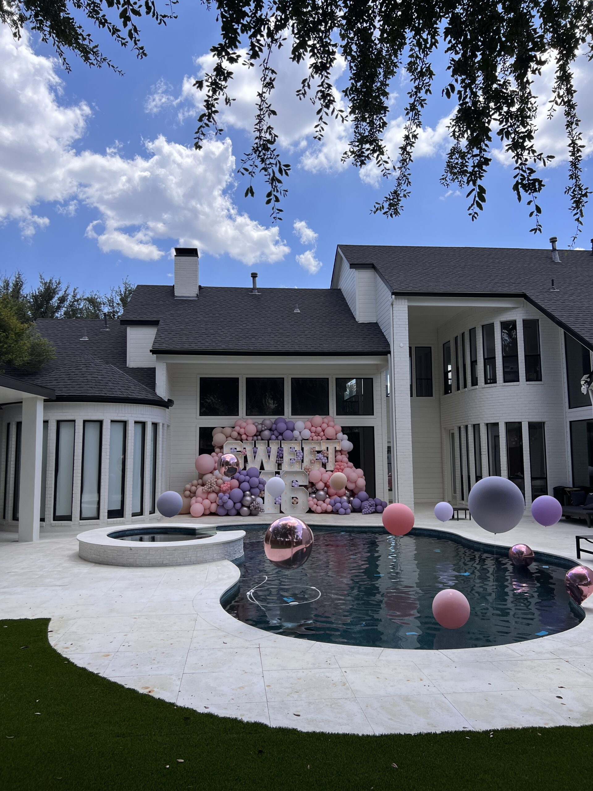 A backyard decorated for a "Sweet 16" celebration features a pool with floating balloons and a colorful balloon arch backdrop.